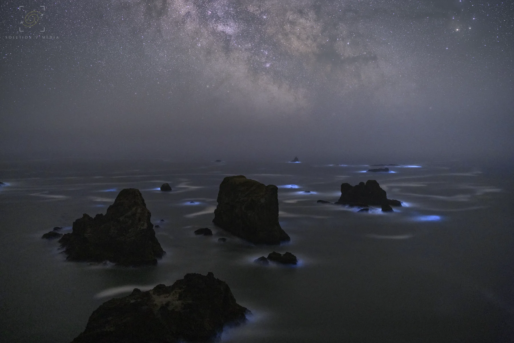 Blue bioluminescence glowing around Pacific coast sea stacks under the Milky Way